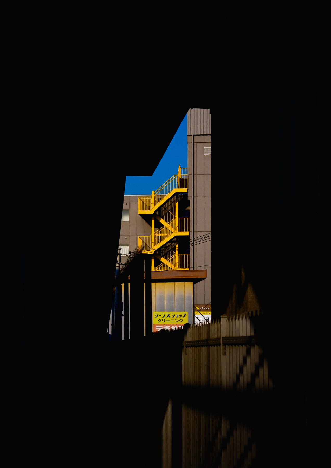 A yellow outdoor staircase beneath a blue sky, framed by deep shadow.